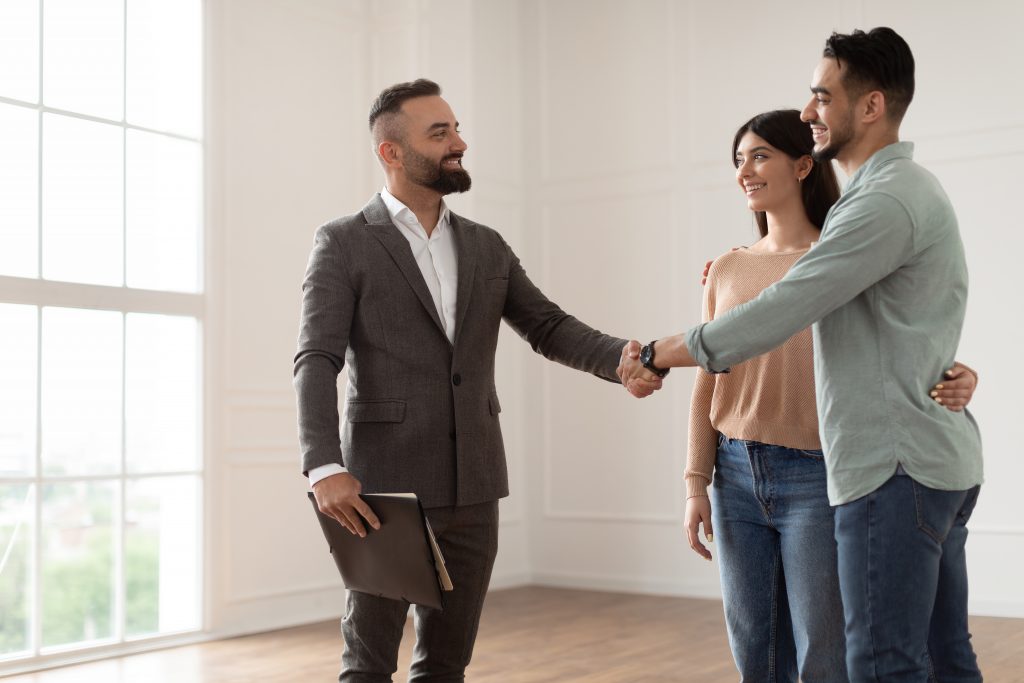 Sealing A Deal Concept. Portrait Of Millennial Couple Buying New Apartment, Shaking Hands With Professional Real Estate Agent. Smiling Manager In Suit Selling Flat To Happy Young Family