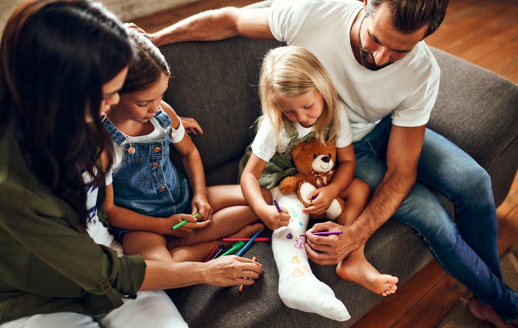 Little girl with a broken leg on the couch. Mom, dad and two daughters draw on plaster with felt-tip pens. The whole family is having fun at home.