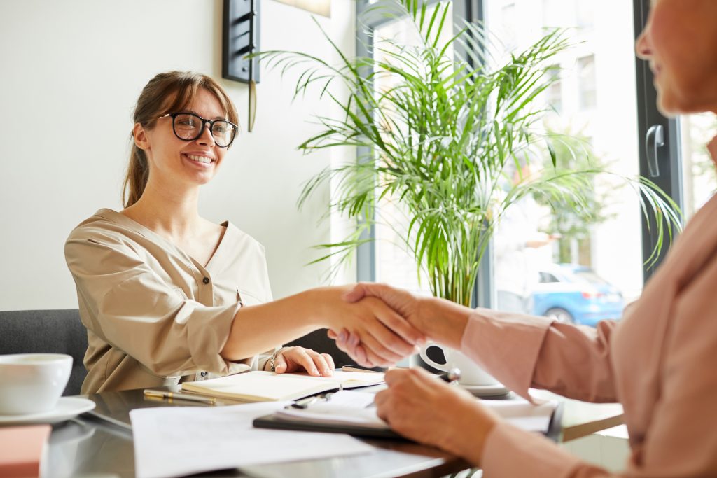 Successful young businesswoman in eyeglasses shaking hands to her partner and smiling while they working at the table at office