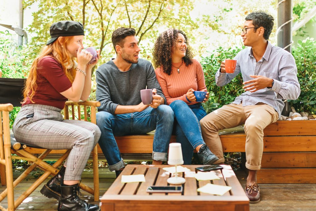 Group of multicultural young friends hanging out on a patio chilling together over mugs of coffee laughing and joking against a high key background