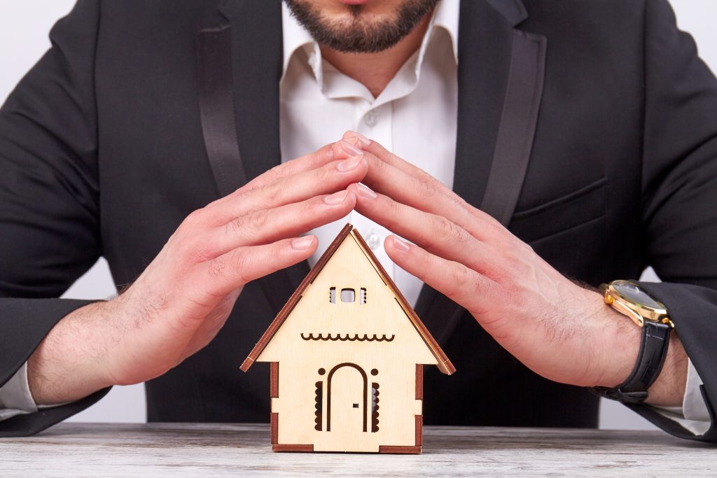 Real estate agent offers you a new house. Businessman in suit protecting house model at office desk. Concept of real estate insurance.