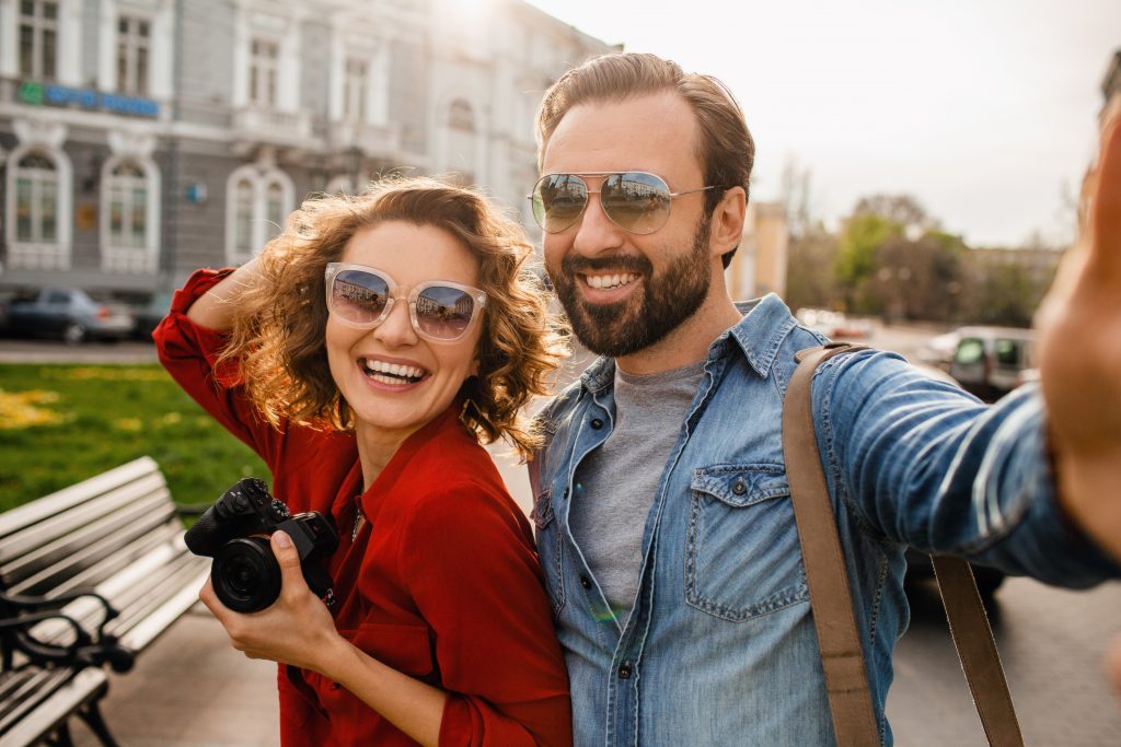 attractive laughing man and woman traveling together, stylish couple in love taking selfie photos on phone on romantic trip, sunny summer city, wearing shirt, sunglasses, travelers having fun