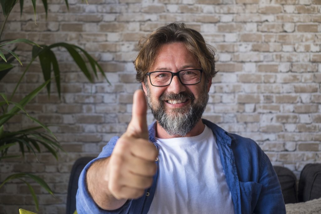 Portrait of cheerful mature man in eyeglasses giving approval or appreciations sign by gesturing thumbs up in front of brick wall. Happy caucasian male looking at camera and giving a signal to proceed