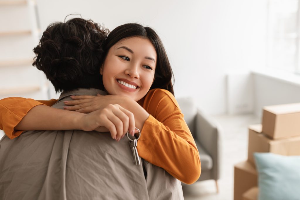 Smiling young Asian woman hugging her boyfriend and holding house key on moving day, free space. Loving millennial couple embracing in their new home during relocation