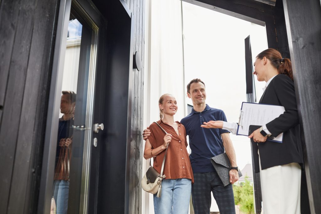 Waist up portrait of female real estate agent meeting young couple standing in doorway while buying new house, copy space