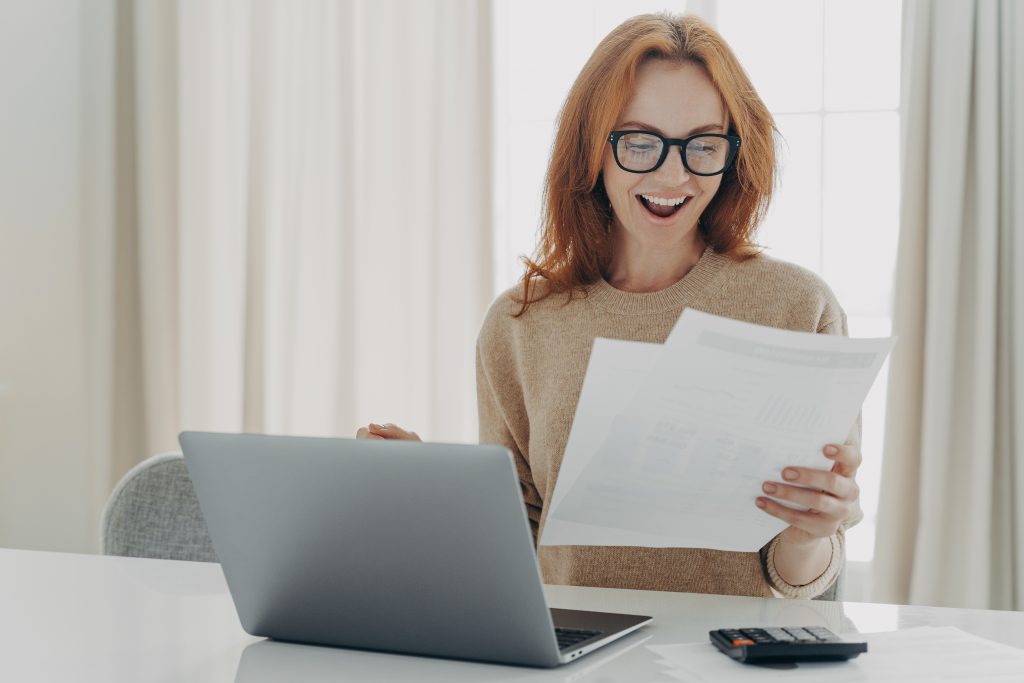 Happy excited redhead woman reading paper letter with notification about last mortgage payment while calculating family monthly budget and paying bills online at home, using laptop and calculator