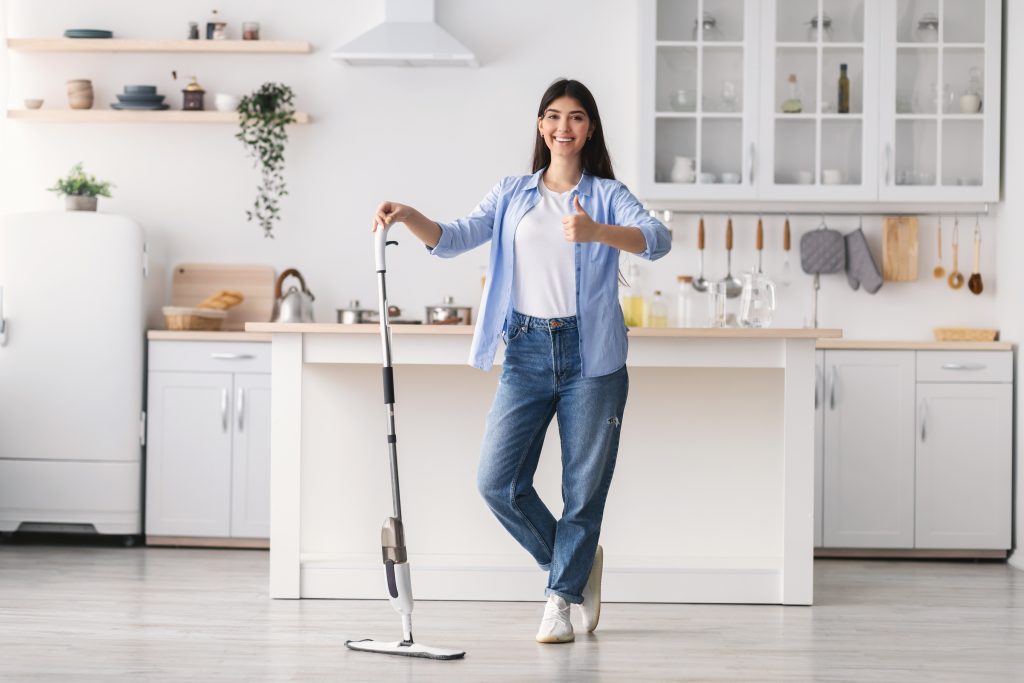 Smiling woman showing thumb up sign gesture after washing hardwood laminate flooring, holding and leaning on water spray mop pad, standing at kitchen posing at camera, full body length