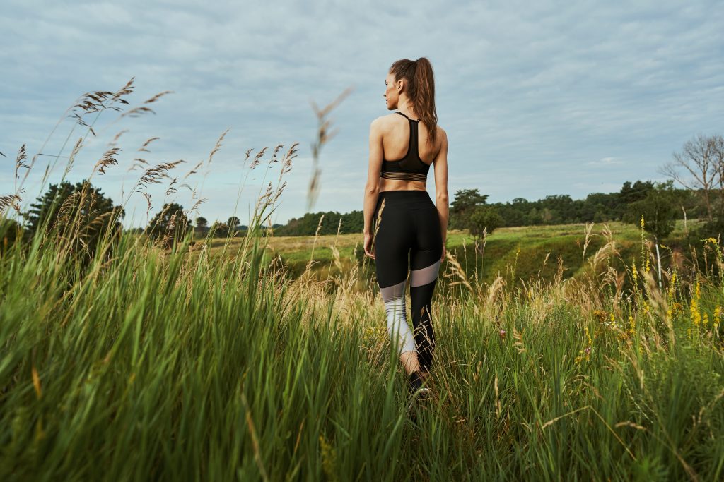 Calm young woman wearing fitness top and sports leggings standing in a beautiful meadow and looking into the distance