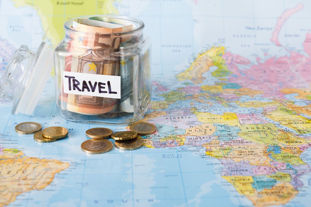 Travel budget concept. Money saved for vacation in glass jar on world map background, copy space. Banknotes and coins for big adventure.