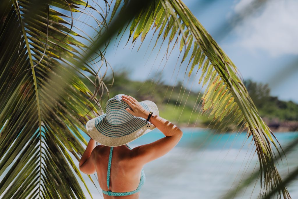 Woman on the beach in the palm trees shadow wearing blue hat. Luxury paradise recreation vacation concept.