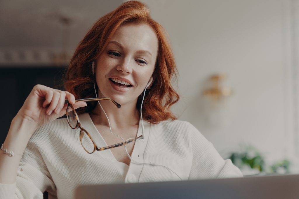 Confident pleased redhead woman concentrated in laptop computer, watches video or movie on laptop computer, enjoys friendly video call, holds transparent glasses, uses free internet connection