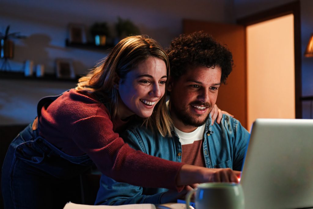Young happy caucasian couple browsing on internet using a laptop to search sale at home. Excited husband and cheerful wife smiling and looking for new apartment sitting on a desk at living room. High quality photo