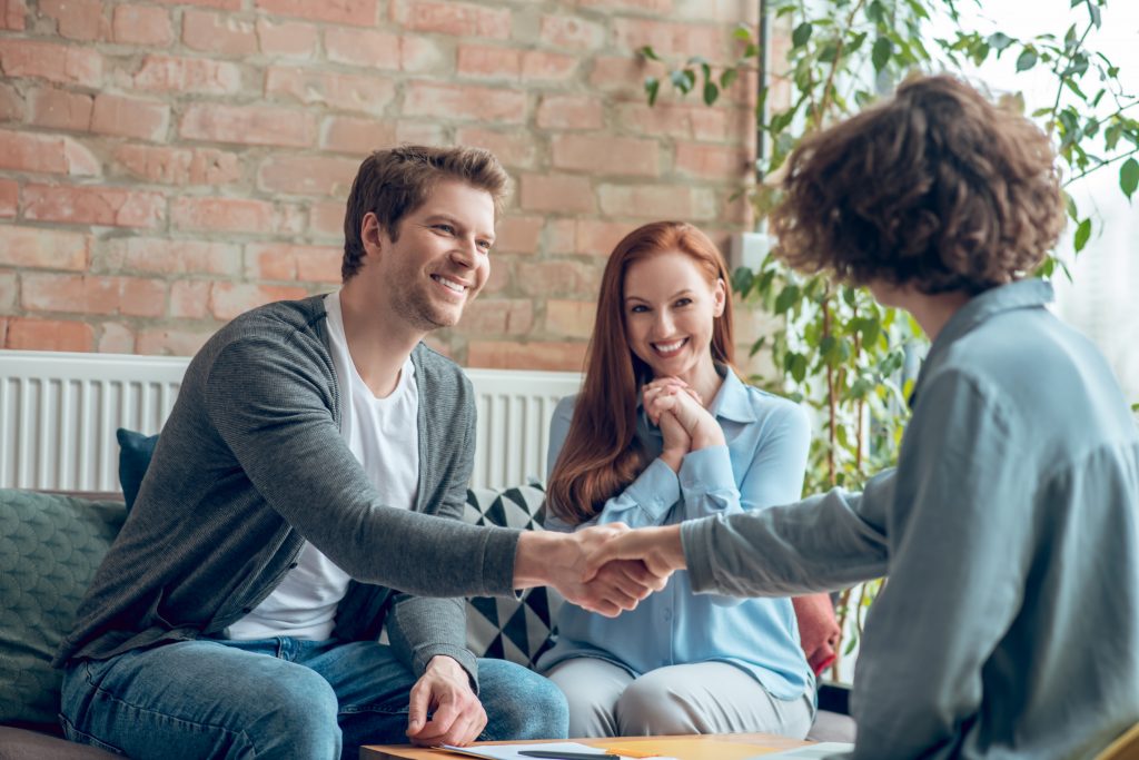 Good moments. Smiling young man and broker shaking hands and happy long haired woman sitting at table in office during afternoon