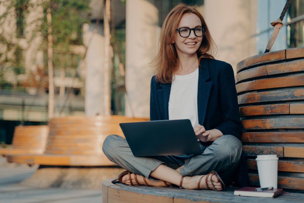 Pleased pretty redhead woman learns educational course on laptop computer sits crossed legs on wooden bench outdoors drinks takeaway coffee looks happily into distance creats new publication