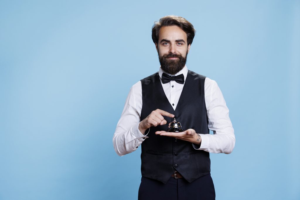 Hotel concierge holds service bell used to ring for assistance and help at front desk. Male bellboy empoyee in formal suit presenting hospitality industry, doorkeeper porter job.