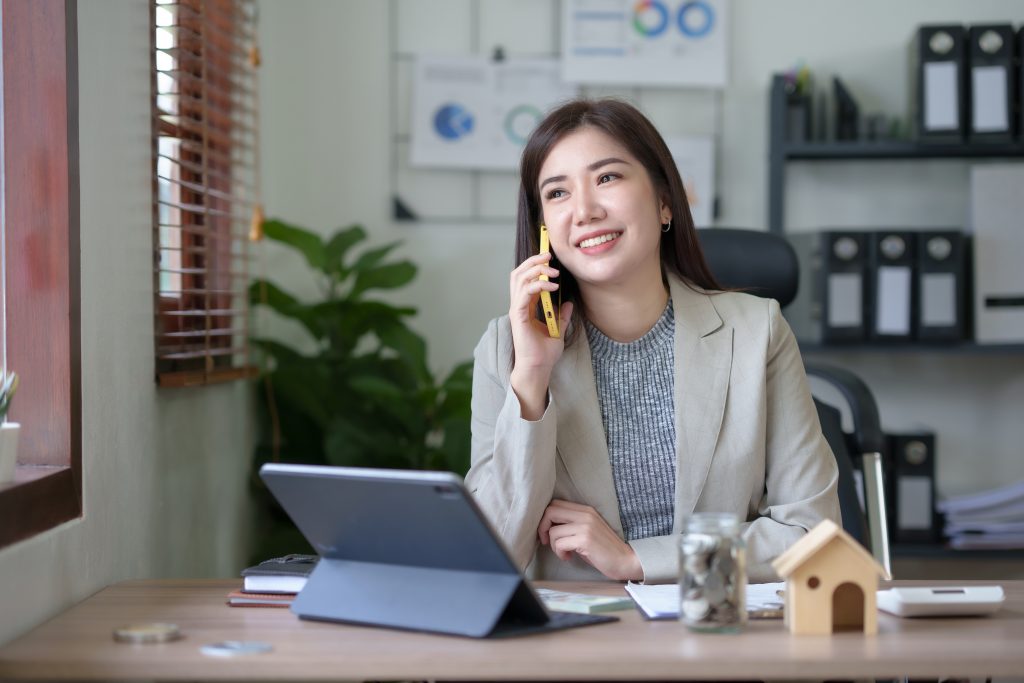 Asian businesswoman, real estate agent, talking on the phone with a customer about the details of selling a house How to buy a house step by step in the office and use the tablet.